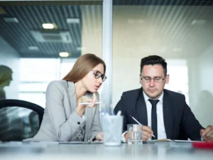 Two people haviing a Two collogues working and seating together in an office setting, holding pens with one holding it up a high and one using the pen to write, serious and discussing work. 