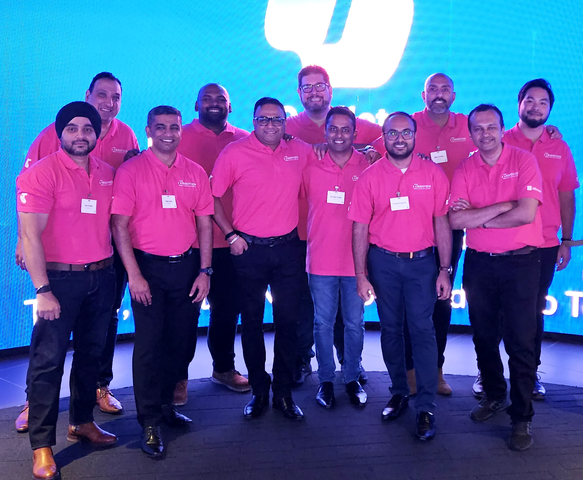 Exigo Tech team members wearing matching pink shirts, posing together at a company event in front of a bright digital display.