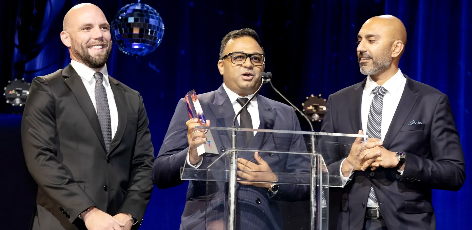 Exigo Tech management team members in formal attire on stage at an awards ceremony, with one speaking into a microphone while holding a trophy.